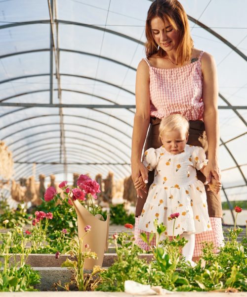Baby is standing. Mother and daughter are in the greenhouse